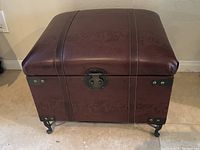 Front view of brown leather ottoman showing the cushioned top, metal clasp, and metal reinforced corners with rivets.