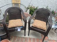 Two brown plastic wicker chairs with patterned cushions placed outdoors on a brick and concrete surface with plants in the background.