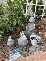 Four rabbit and bunny garden figurines arranged outdoors on rocks and mulch near green shrubbery and a lattice.