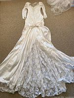 Full view of the wedding dress laid flat showing satin fabric with lace and floral filigree embellishments, puff sleeves, bow detail, and long lace train extending behind.