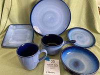 Display of various pieces including dinner plates, square plates, mugs, onion bowls, and smaller plates arranged on fabric.