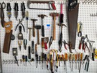Wide view showing assortment of hand tools hung on pegboard including saws, hammers, putty knives, pliers, wire cutters, scissors, screwdrivers and more.