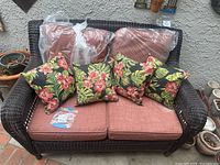 Brown wicker patio sofa with red-orange seat cushions and four tropical floral decorative pillows. Three red-orange cushions in plastic laid out on sofa back.