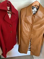 Red wool Cabi coat and tan polyurethane Cabi jacket hanging side by side on a wooden rod.