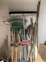 Overview photo showing large pushbroom, various rakes including Flex Rake, gardening tools like shovels and hoes, and other long handled tools stored upright against a wall.
