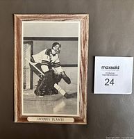 Front view of the Jacques Plante hockey photo mounted on cardboard showing the player in goal with a woodgrain border and name plate.