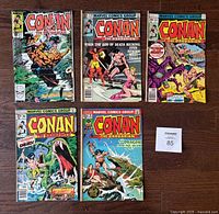 Top view of 5 Conan the Barbarian Marvel comics arranged on a wooden floor, showing front covers with various action scenes and characters.