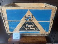 Front view of the Pure Spring wooden beverage crate showing blue and beige painted logo with crown and triangle design.