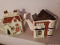 Two vintage porcelain teapots shaped like houses viewed from the front, showing their spouts and detailed designs.