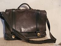Front view of a medium-sized brown leather satchel showing the flap, shoulder strap, and top handle. Two gold-colored turn-lock clasps secure the flap.
