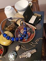 Clear view of multiple vintage and curiosity items including blue beaded necklace, wristwatch, ceramic mugs, brass shoe decoration, small ceramic bust, coins or medallions, and small leather key holder.