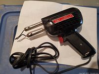 Black electric soldering gun with pistol grip and red trigger, lying on a white surface with the cord nearby.