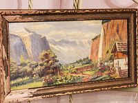Full view of vintage oil painting showing landscape scene with cliffs, house, and vegetation in worn wooden frame.