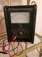 Front view of vintage Simpson voltage meter tester showing main dial, handles, selector knob and attached test leads.
