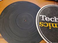 Top view of black rubber circular record pad with printed Technics logo in white and yellow on black background, showing surface detail and wear.