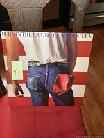 Front cover of Bruce Springsteen Born in the USA vinyl album showing man from behind holding a red cap, dressed in jeans and white t-shirt against red and white striped background.