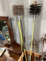 Two chimney brush heads attached to yellow extension poles leaning against a wall, with additional poles stacked nearby.