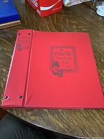 Two red binders on a wooden surface featuring the Coca-Cola Collectors Club Int'l black embossed design on front and spine.