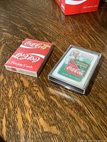 Photo showing two decks of Coca-Cola playing cards on wooden surface, one red box unopened, the other in clear plastic case