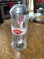 Full view of glass Coca Cola straw dispenser with metal lid on wooden table.