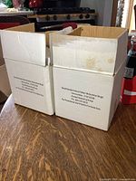 Photo showing two cardboard boxes labeled 'Small Shaded Votive Holder-Midsummer Magic' with promotional text and item number 5132-CC39 indicating the product packaging.