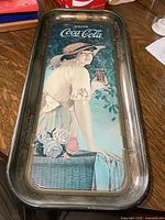Photo of rectangular metal Coca Cola tray showing a woman in a hat holding a glass of Coke, with floral and basket details.