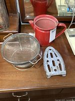 Photo of all three items placed together on a wooden surface: the red enamel pitcher, metal mesh strainer, and cast iron trivet.