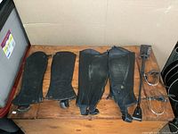 Two pairs of black leather half chaps, metal riding spurs with black leather straps, and a new black riding crop with tag laid on wooden surface.