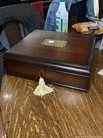 Closed dark wood jewelry/storage box with brass plate on top and gold-colored tassel on key inserted into lock on front side.