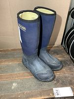 Pair of dark blue tall boots with black rubber foot section and dark blue waterproof upper, displayed standing on wooden surface against cardboard background.