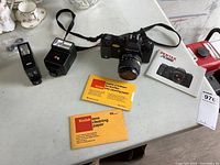 Photo of Pentax A 3000 35mm film camera with attached Pentax-A Zoom 35-70mm f/2.8 lens, black strap, Braun flash, camera manual, and Kodak lens cleaning paper on a white surface.