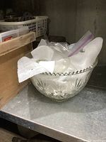 Clear glass punch bowl partially filled with white packing materials and plastic ladle inside, showing scalloped edge and starburst pattern.