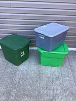 Three large storage totes: grey Sterilite latching box on top of two green totes against siding background.