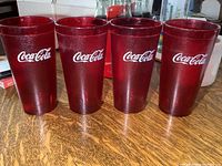 Four red translucent plastic Coca-Cola cups shown standing upright on a wood surface.