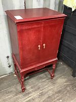 Front and side view of the red vintage radio cabinet with two doors and wooden knob handles standing on turned legs.