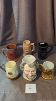 Seven steins and mugs arranged on dark cloth with gray backdrop, showing various sizes and styles.