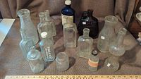 Wide view of twelve assorted antique glass bottles arranged on fabric surface showing shapes, sizes, colors and embossed markings.