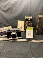 Minolta Hi-Matic 35mm camera and Kodak Folding Autographic Brownie camera with their respective manuals and cases displayed with ruler