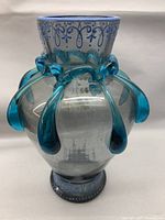 Ornate clear glass vase with blue glass drip accents around the top and handpainted floral designs on the body, standing on a flat surface