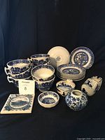 Lot composed of blue and white china including eight tea cups with Willow pattern, eight matching saucers, a trinket dish, commemorative coaster, Dutch clog shaped ashtray, small Thailand vase, and a heart-shaped trinket box with lid. All items arranged on black background.