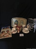 Display of tea cups with saucers, mugs, tea tin, commemorative serving tray, and collector magazines.