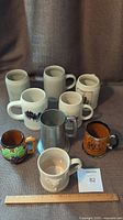 Photo showing all 9 steins and mugs of various sizes and materials grouped together with a ruler for scale.