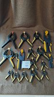 Full collection of 16 clamps arranged on a brown cloth background showing the size and types of clamps included with black and yellow rubberized handles.