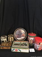Seven vintage collectible tins arranged on a table with black backdrop, clearly showing a range including Coca-Cola, Peek Frean, and decorative older tins.