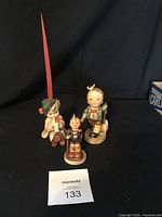 Photo showing three Goebel Hummel figurines displayed on a black background with auction lot number card visible.