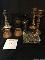 Seven candlesticks including a tall silver-tone Art Deco style, two black etched brass lacquered enamel, two brass fluted base, and two small glass square holders arranged on black background.