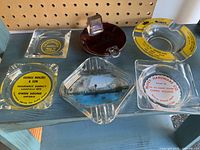 All six ashtrays displayed on blue table surface with pegboard in background. Includes antique red Owen Sound ashtray, clear glass ashtrays with yellow business advertisements, and image of S. S. Norie ship. Visible chip on King Insulation ashtray and soiling near ship image.