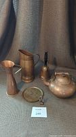 Photo showing kettle, large pitcher, small pitcher, bell-shaped money bank, and copper kettle on grey textured fabric background.