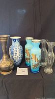 Full view of all six vases: two blue koi fish ceramic vases with gold rims, ceramic blue flower vine vase, brass engraved vase, and two clear glass cornflower vases.