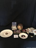 Full lot showing all commemorative plates, metal tins, handkerchief, and ashtray arranged against black backdrop